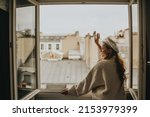 Small photo of A woman stands near the window against the backdrop of the roofs of Paris. A girl with long hair in a beige suit and a beret enjoys the city and the trip waving to her neighbors.