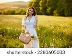 a beautiful woman in a light dress and a basket of daisies in her hands stands in a field during sunset and smiles at the camera