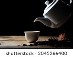 Hot tea is pouring from a teapot into a white cup with herbs on a black background