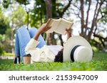 Side view of charming woman reading book in grass under tree with cup of coffee. Relax in summer time holiday laying on the grass field comfortable feeling.