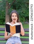 Small photo of Surprised young woman with widely open yeas and mouth and a hand on cheek is reading a book outdoors.