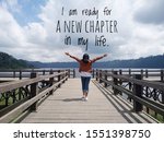 Inspirational motivational quote - I am ready for a new chapter in my life. With young woman standing on wooden bridge, hands raised with open arms against the blue sky & lake view background.