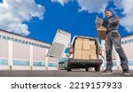 Small photo of Postal worker. Minivan behind courier. Postman picks up boxes from warehouse. Postman in gray uniform. Postal delivery worker. Career in courier company. Postman with clipboard near warehouse