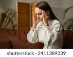 Small photo of A tired woman with a severe headache. A young girl in a state of stress, suffering from acute migraine or pain with temporal arteritis, is sitting at home on the couch.
