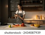 Small photo of A young woman dressed in casual attire with a grey apron is sitting in a well-lit modern kitchen. She is focused on her smartphone, perhaps following a recipe or chatting, surrounded by fresh
