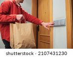 Small photo of Delivery man in protective mask holding paper bag with food in the entrance. The courier stands at the door and rings the customer's doorbell. He brought a box of fresh vegetables and fruits.