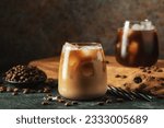 Ice coffee in a tall glass with cream poured over, ice cubes and beans on a dark concrete table. Cold summer drink with tubes on a black background with copy space.