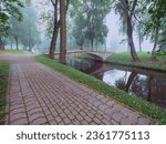 Small photo of Brick foot path to a small concrete old bridge over a small river in a town forest park in a fog. Mistry surreal calm mood. Relaxing atmosphere and melancholic nature vibe.