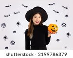 Halloween theme, young asian pretty woman in black halloween costume wearing witch hat holding orange pumpkin posing smiling on black spider and but on white background.