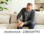 Small photo of Knee pain arthritis body sick health care concept. Unhappy middle aged senior man suffering from knee ache sitting on sofa at home. Mature old senior grandfather touching leg feeling pain hurt in knee