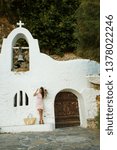 Small photo of CRETE, GREECE - SEPTEMBER 15, 2018: A beautiful tanned brunette girl in a tight-fitting pink dress stands next to a small Greek church.