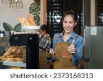 The owner of small cafe is beautiful Asian business woman. Standing behind the bakery phone in hand, gave thumbs up smile. There  baguette in the laundry basket. Retired mother making coffee the back.