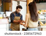 Small photo of Indian male coffee shop owner young businessman serves brown disposable paper coffee cup Asian customer woman behind cash register smilelips head slightly bowed bakery cabinet cafe small business.