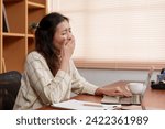 Small photo of Yawning from fatigue, an Asian office worker takes a brief respite from her laptop work to refresh. overworked Asian professional caught mid-yawn, signaling a long day in the home office