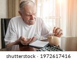 Small photo of old man in the pandemic counts pennies from the pension fund. Concerned about lockdown, an elderly man puts money in a piggy bank. pensioner calculates the budget of his pension.