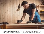 Experienced carpenter in work clothes and small business owner working in woodwork workshop,  using sandpaper for polishing  wooden bar  at worktable in workshop  