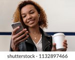 The girl holds a white glass in her hand, looks into the phone and smiles. A beautiful young modern black woman, in a leather jacket with airpods in her ear.
