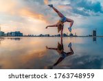 Small photo of Female gymnast standing on one hand and keeping balance during dramatic sunset with reflection in the water of amazing clouds. Concept of Calisthenic, contortion and handstand