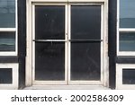 Double commercial exterior doors of a store. The glass has been painted black, trim is black and beige. The old doors have metal handles. There are large glass windows on both sides of the doors. 