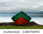 Bright orange and green colorful wooden row boat on a beach. There is a blue ocean, large trees covering a mountain with fog in the background. Green grass on the ground in front of the boat.