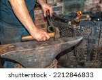 Small photo of A forger wearing blue jeans and a t-shirt holds fire thongs and a metal twisted bolt on a vintage anvil. There's rounding hammer tool with a wooden handle in the blacksmith foundry shop with a furnace
