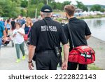Small photo of Medical first responders walk along a road wearing black uniforms, with the medical first responder in grey letters across the back of the paramedic. The EMT is carrying a red first aid kit and radio.