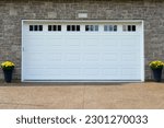 Small photo of A large white metal panel residential garage door with small glass windows on a grey brick building. There are two pots with yellow flowers on either side of the door. The ground is a beige block.
