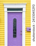 Small photo of The entrance to a vibrant yellow wooden house with clapboard siding and a purple mailbox. There's a single purple metal door with a narrow glass decorative window, white trim and a transom window.