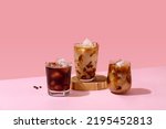 Ice coffee in a tall glass with cream poured over and coffee beans. Set with different types of coffee drinks on a pink table.