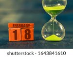 Small photo of february 18th. Day 18 of month,Handmade wood cube with date month and day and hourglass with green sand. Time passing away. artistic coloring. winter month, day of the year concept