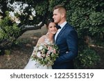 Beautiful newlyweds cuddle in a green garden with trees. Young bride and cute bride with a bouquet are standing in the park. Wedding photography. Stylish portrait.