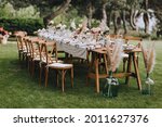 A large, long, decorated, wooden table and chairs, covered with a white tablecloth with dishes, flowers, candles, stands on the green grass in the park, in the forest in nature. Wedding banquet.