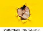 Small photo of Close-up of a woman's ear and hand through a torn hole in the paper. Yellow background, copy space. The concept of eavesdropping, espionage, gossip and tabloids.