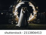A stylish groom and a beautiful bride in a long dress are standing, hugging at night near a luminous cane arch decorated with lamps and garlands. Wedding photography, portrait.