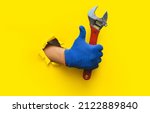Small photo of The right hand in a blue knitted glove holds a red wrench and gives a thumbs up (like). Torn hole in yellow paper. The concept of a worker, a labor migrant, a master of his craft. Copy space.