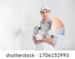 Painter or decorator with handful of colorful paint swatches or color cards holding a roller in his other hand as he smiles at the camera against a white wall