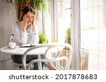 Small photo of young tired woman in depression with a headache sits in a modern cafe holding her head with her hands. Joyless unmotivated employee at lunch in a restaurant. Spasm of the vessels of the head.