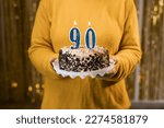 Small photo of Happy ninetieth birthday. Woman holding fresh delicious birthday cake with burning candle number 90, close up. Celebration of birthday at home. Copy space