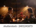 Small photo of Happy young couple celebrating Valentines day having romantic dinner at home. Loving man giving gift box his woman. Copy space