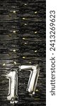 Small photo of Silver foil balloon number 17 on a background of black tinsel decoration with golden confetti. Birthday greeting card, inscription seventeen. Anniversary event. Vertical Banner. copy space.