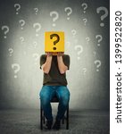 Small photo of Incognito young man seated on a chair holding a yellow box with question mark instead of head. Introvert person anonymity concept hiding identity behind a mask. Social issue, shy guy covering face.