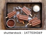 Homemade chocolate popsicles on ice in rustic tray on wooden background. Chocolate fudgesicles with chocolate and coconut flakes - horizontal image 