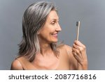 Small photo of Senior woman with gray silver hair holding eco toothbrush and looking at it isolated on grey. Beautiful mature lady in beige undershirt going to cleaning the teeth with bamboo brush. Dental care