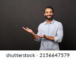 Small photo of Happy Indian bearded man in casual shirt points hands aside at empty space, hispanic man advertising novelty standing isolated on black, showing with palms sideway