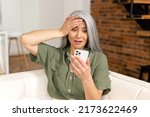 Small photo of Worried senior woman staring at a smartphone screen at home, upset retire female sitting on the couch looks at mobile phone with vexation, read terrible news or received bad analysis results