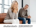 Small photo of Upset middle-aged couple is arguing at home, frustrated blonde woman covered face with palms and crying, sadness grey-haired mature man on background. Spouses have difficulties, relationship crisis