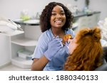Small photo of Smiling black woman dentist wearing blue uniform, providing tooth restoration and filling with curing polymerization UV lamp for her little patient, cute girl with red curly hair