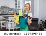 Smiling female cleaner in green apron showing thumb up and holding bucket with with detergents and rags. Cleaning service and housekeeping concept.