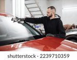 Car detailing and cleaning concept. Good-looking young male car wash worker, wearing black gloves, wipes and polishes car windshield window with microfiber cloth.
