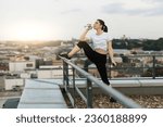 Small photo of Young fit lady in yoga outfit drinking beverage while leaning with right leg on handrails of building roof. Beautiful adult restoring fluid balance after strenuous athletic activity in fresh air.